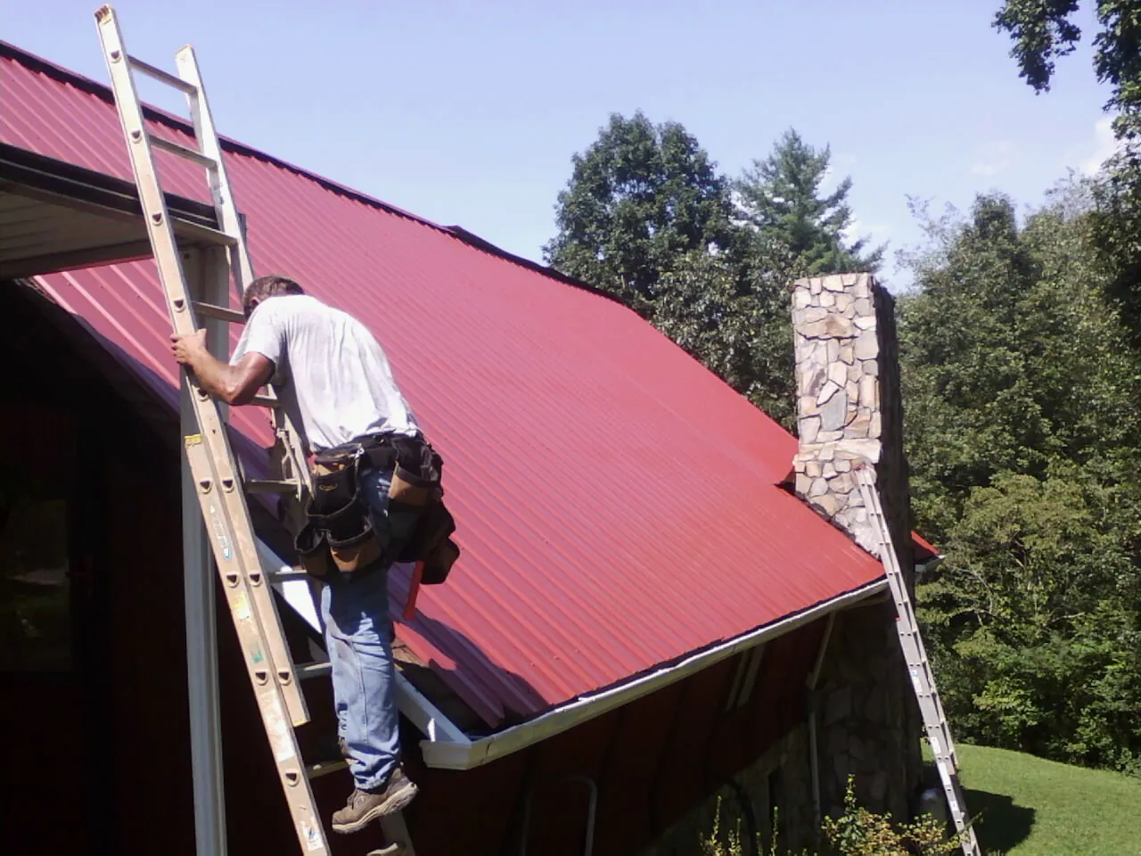 Roofer on a ladder beside a red metal roof and stone chimney — Soffit & Fascia Repair by Roofing Brothers in Hatboro